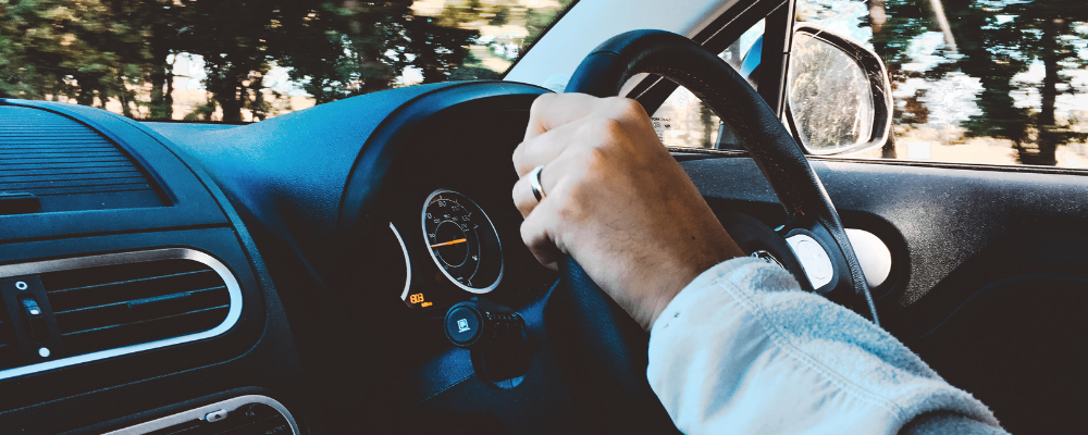 A hand wearing a ring grips the steering wheel of a car, with the dashboard and a side mirror visible. Trees blur past outside the window, indicating movement.
